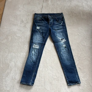 Blå slitna jeans från Dsquared2 - Säljer ett par blå jeans från Dsquared2 med slitningar och färgstänk. Byxorna har fem fickor, knappgylf och en röd Dsquared2-logga vid gylfen. Passformen är rak och materialet är klassisk denim.