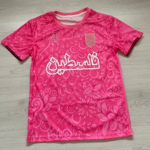 Rosa fotbollströja med blommigt mönster och arabisk text - Säljer en rosa fotbollströja med blommigt mönster och vit arabisk text på bröstet. Tröjan har korta ärmar och en rund halsringning med rosa kant. På vänster bröst finns en sköld i guld och rosa. Perfekt för dig som vill sticka ut på planen.