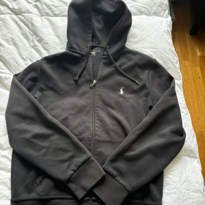Svart hoodie med dragkedja från Ralph Lauren - Säljer en svart hoodie från Ralph Lauren med dragkedja framtill och klassisk broderad logga på bröstet. Tröjan har huva med snörning och långa ärmar. Perfekt för en avslappnad stil.