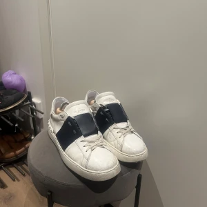 Valentino Open Marinblå - Säljer ett par vita Valentino Open sneakers med bred mörkblå rem över mitten. Skorna har rund tå, snörning och klassisk platt sula. Ikoniska nitar baktill på sulan och diskret Valentino-logga på plösen. Medföljer box och dustbags. Har även äkthetsbevis. Storlek: 46 