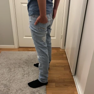 Ljusblå jeans med feta slitningar  - Säljer ett par ljusblå jeans med slitningar och trasiga detaljer framtill. Jeansen har raka ben, klassiska fem fickor och orange sömmar. Perfekta för en avslappnad och trendig stil. Perfket för kyligare sommarnätter och passar till alla olika sorters outfits. ”Låga i midjan”Nypris 7000+. Vårt pris 899. Riktigt steal