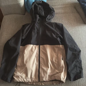 Svart och beige vindjacka från Didriksons - Säljer en svart och beige vindjacka från Didriksons med huva och dragkedja framtill. Jackan är 100% vattentät och har stor logga på ryggen. Perfekt för blåsiga och regniga dagar. Snygg tvåfärgad design och praktiska fickor.