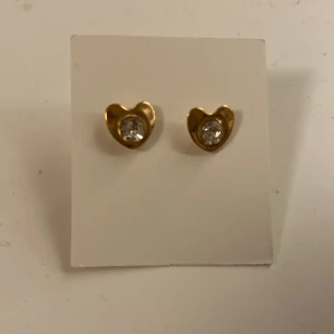Guldiga hjärtformade örhängen med sten - Säljer ett par söta örhängen i guldton, formade som hjärtan med en glittrande klar sten i mitten. Perfekta för att ge din look lite extra shine och romantisk touch.