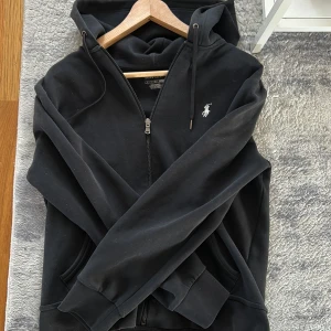 Svart hoodie med dragkedja från Ralph Lauren - Säljer en svart hoodie från Ralph Lauren med dragkedja och huva. Tröjan har två fickor framtill och den klassiska loggan broderad på bröstet. Perfekt för en avslappnad stil. Storlek S