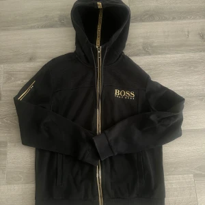 Hugo Boss hoodie  - Svart hoodiejacka från Hugo Boss med guldfärgade detaljer och dragkedja. Köpt på Zalando för ett par år sedan. Nypris 1800kr
