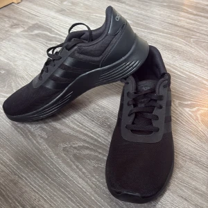 Svarta Adidas sneakers - Säljer ett par helsvarta Adidas sneakers med klassiska tre ränder på sidan. Skorna har snörning, platt sula och är tillverkade i syntetmaterial och mesh för bra andningsförmåga. Perfekta för vardagsbruk eller träning.