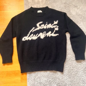 Svart stickad tröja från Saint Laurent - Säljer en svart stickad tröja från Saint Laurent med vit text på bröstet. Tröjan har rund halsringning och ribbade muddar vid ärmslut och nederkant. Perfekt för dig som gillar exklusiva och stilrena plagg. Aldrig använt. Hör av dig vid minsta fundering!!