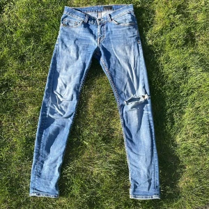 Tiger of Sweden ljusblå jeans Evolve - Säljer ett par ljusblå jeans från Tiger of Sweden, modell Evolve. Jeansen har klassisk femficksdesign, slitningar och hål på ena benet för en trendig look. Tillverkade i Italien med snygga kontrastsömmar och normal passform.