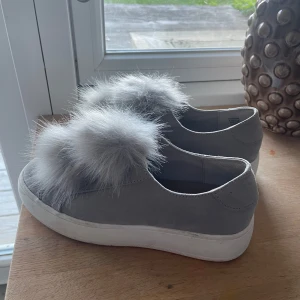 Grå sneakers med pälsdetalj - Säljer ett par grå sneakers med vit sula och stora fluffiga pälsdetaljer ovanpå. Skorna har en rund tå och är i mockaliknande material. Från Steve madden i storlek 37