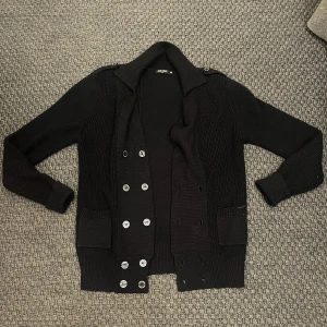 Svart stickad kofta från Antony Morato - Säljer en svart stickad kofta från Antony Morato med dubbelknäppning framtill och två fickor. Koftan har långa ärmar och en klassisk krage. Perfekt för lager-på-lager och stilrena outfits. Väldigt grisch!