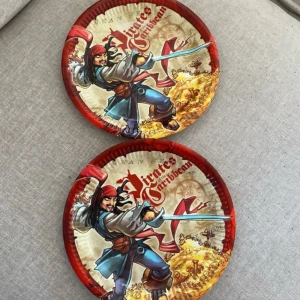 Pirates of the Caribbean papptallrikar oanvända  - Två runda papptallrikar med motiv från Pirates of the Caribbean. Tallrikarna har en färgglad design med en tecknad pirat, skattkarta och svärd. Perfekta för temakalas eller barnkalas med pirattema.