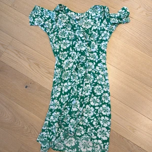 Grön blommig klänning från Monki - Säljer en grön kortklänning från Monki med vitt blommigt mönster. Klänningen har korta ärmar och en lätt v-ringning. Perfekt för varma dagar och har en luftig passform.