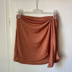 Rostfärgad omlottkjol - Säljer en kort rostfärgad kjol med omlottdetalj och drapering framtill. Kjolen har en mjuk och följsam känsla.