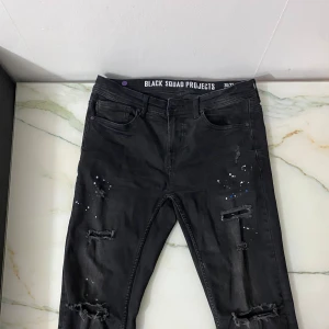 Svarta jeans från Black Squad Projects - Snygga svarta jeans med slitna detaljer och färgstänk från Black Squad Projects. De har en normal passform och är perfekta för en avslappnad stil. Byxorna har fem fickor och en knappgylf.