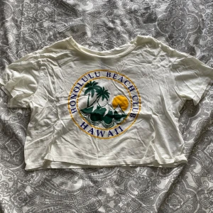 Vit croppad t-shirt från H&M Divided med tryck - Säljer en vit croppad t-shirt från H&M Divided med färgglatt tryck framtill: 'Honolulu Beach Club Hawaii' med palmer, sol och vågor. Perfekt för sommaren och strandhäng. Mjuk bomullskvalitet och kortärmad modell.