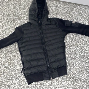Svart pufferjacka med huva från Moncler - Säljer en svart pufferjacka från Moncler med huva och dragkedja framtill. Jackan har quiltad kropp och släta ärmar, samt ribbade muddar vid ärmslut och nederkant. Perfekt för kyliga dagar.