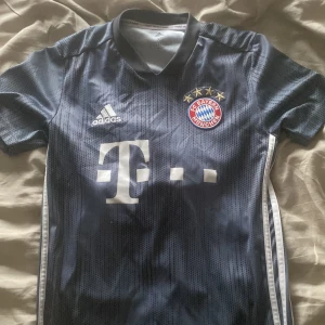 Bayern München bortatröja från Adidas - Säljer en mörkblå Bayern München bortatröja från Adidas med vita detaljer och klubbmärke på bröstet. Tröjan har korta ärmar, V-ringning och klassiska Adidas-ränder längs sidorna. Perfekt för fotbollsfans!
