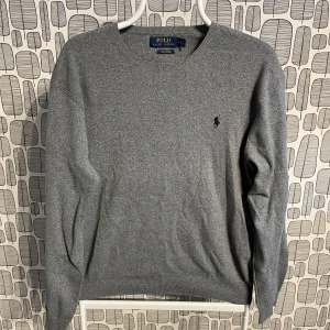 Grå tröja från Polo Ralph Lauren - Snygg grå tröja från Polo Ralph Lauren i mjuk pima bomull. Den har en klassisk rund hals och ett diskret broderat märke på bröstet. Perfekt för en stilren look.
