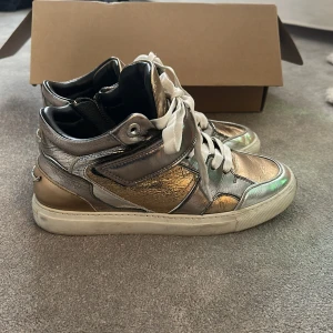 Metallic sneakers från Zadig & Voltaire - Snygga metallic sneakers från Zadig & Voltaire i silver och guld. Skorna har snörning och dragkedja på sidan för enkel på- och avtagning. Perfekta för att ge din outfit en cool touch. Box och dust bag kommer med, vill man ha fler bilder är det bara att fråga. Nypris 4000kr