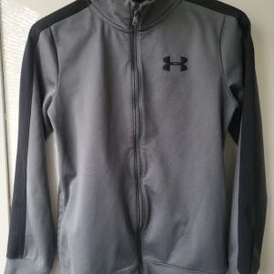 Grå träningsjacka från Under Armour - Säljer en grå träningsjacka från Under Armour med svarta detaljer. Jackan har en dragkedja framtill och långa ärmar. Perfekt för träning eller som en lätt överdragströja. Den har en stilren design med logotyp på bröstet.