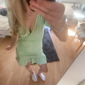 Grön rutig omlottklänning - Söt grön rutig omlottklänning med volangdetaljer. Klänningen har en v-ringad design och korta ärmar, perfekt för sommarens varma dagar.