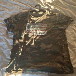 Camouflage t-shirt från Ralph Lauren - Säljer en cool camouflage t-shirt från Ralph Lauren med ett slitet amerikanskt flaggmotiv på bröstet. T-shirten är kortärmad och tillverkad i bomull, perfekt för en avslappnad stil.