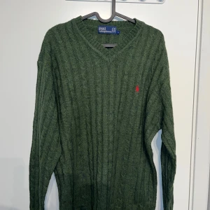 Grön stickad tröja från Ralph Lauren - Säljer en snygg grön stickad tröja från Ralph Lauren med v-ringning och det klassiska röda logotypbroderiet på bröstet. Perfekt för kyliga dagar och ger en stilren look. Passar både till jeans och chinos.