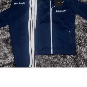 Mörkblå dress från Palm Angels - Säljer en snygg mörkblå dress från Palm Angels med vita detaljer. Jackan har dragkedja framtill och vita ränder längs ärmarna. Perfekt för en sportig look eller avslappnad stil. Passar bra till både träning och vardag. 