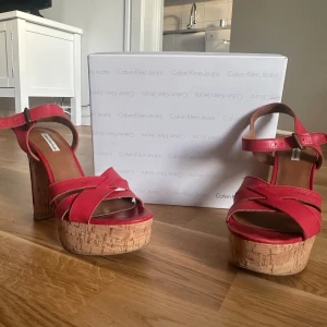 Röda sandaler med korkklack från Calvin Klein - Snygga röda sandaler från Calvin Klein med en hög korkklack och träsula. Skorna har ett korsat remdesign och justerbart spänne vid ankeln❤️