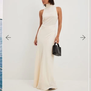 Ljusgul/beige långklänning/ balklänning - Elegant champagnefärgad långklänning i satin med hög hals och öppen rygg. Klänningen är ärmlös och har en vacker knytning i nacken som ger en extra touch av stil. Perfekt för speciella tillfällen.