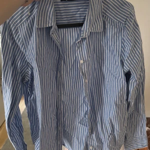 Blå och vit randig skjorta - Säljer en stilren blå och vit randig skjorta med knappar framtill. Skjortan har en klassisk krage och långa ärmar, perfekt för en avslappnad men ändå uppklädd look.