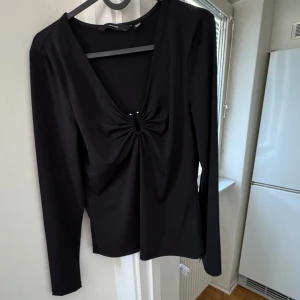 Svart långärmad topp från Vero Moda - Snygg svart långärmad topp från Vero Moda med en elegant v-ringning och knytdetalj framtill. Perfekt för en stilren look. Tillverkad i ett mjukt och bekvämt material.
