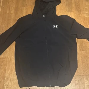 Svart jacka från Under Armour - Säljer en svart jacka från Under Armour med dragkedja och huva. Jackan har en stilren design med Under Armour-loggan på bröstet. Perfekt för träning eller vardagsbruk.