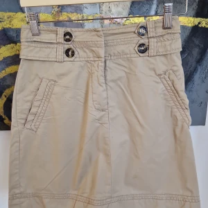 Beige kjol från H&M - Snygg beige kjol från H&M med dekorativa knappar i midjan och praktiska fickor. Perfekt för en stilren look. Den har en klassisk design med en bekväm passform. 47 cm