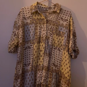 Mönstrad skjorta från Cotton On - Säljer en unik mönstrad skjorta från Cotton On i gult och blåa toner. Skjortan har ett patchwork-liknande mönster med paisley och blommor. Den är kortärmad och har knappar framtill. Perfekt för en avslappnad stil!