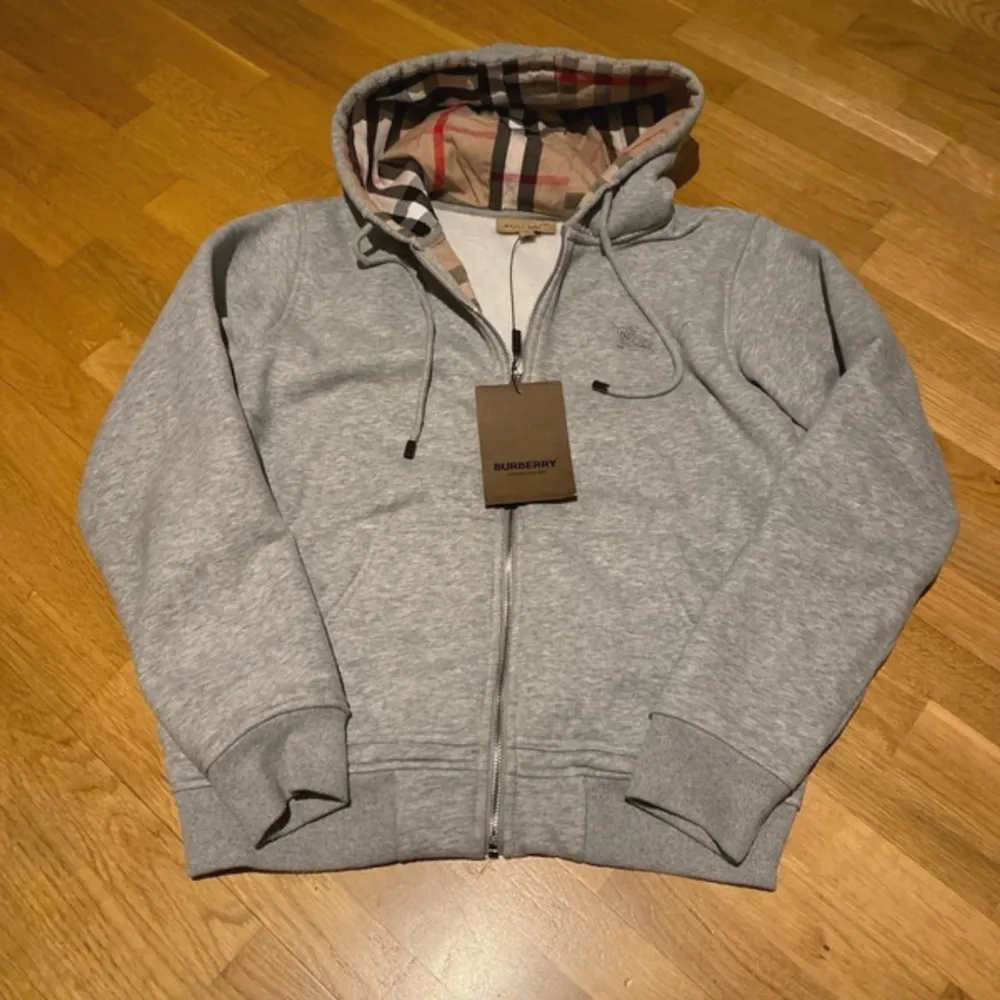 Säljer en stilren grå hoodie från Burberry med klassiskt rutigt mönster i huvan. Tröjan har dragkedja framtill och ribbade muddar vid ärmslut och nederkant. Perfekt för en avslappnad och trendig look.. Neuletakit & Villapaidat.