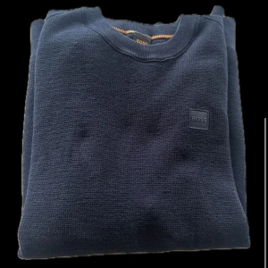 Mörkblå tröja från Boss - Säljer en stilren mörkblå tröja från Boss. Tröjan har en diskret logotyp på bröstet och är tillverkad i ett bekvämt material med ribbade muddar vid halsen. Perfekt för en avslappnad look.