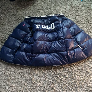 Blå pufferjacka från Ralph Lauren - Säljer en blå pufferjacka från Ralph Lauren med stor POLO-text på ryggen. Jackan har en broderad logga på framsidan och är perfekt för kyliga dagar. Den är långärmad och har en klassisk pufferdesign.