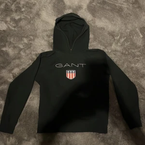 Svart hoodie från GANT - Snygg svart hoodie från GANT med deras logga och ett emblem på bröstet. Tröjan har en klassisk design med huva och långa ärmar, perfekt för en avslappnad stil. Storlek 158-164.