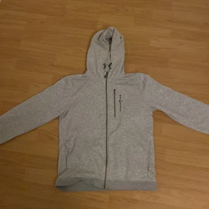 Grå Sail Racing hoodie med dragkedja - Säljer en stilren grå hoodie med dragkedja och huva. Perfekt för en avslappnad look. Den har en diskret logga på bröstet och är tillverkad i ett mjukt material.