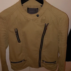 Beige jacka med dragkedjor - Snygg beige jacka från Mango med asymmetrisk dragkedja framtill och quiltade detaljer på axlarna. Jackan har flera dragkedjefickor och en modern design som passar perfekt för höst och vår.