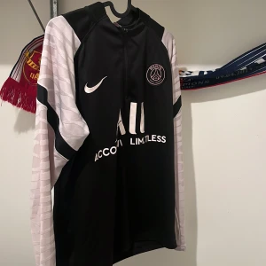 PSG Tracksuit  -  PSG Tracksuit/Säljer en svart fotbollströja med rosa detaljer och matchande byxor från Nike. Tröjan har långa ärmar och PSG-logga på bröstet. Byxorna har elastisk midja och rosa detaljer. Perfekt för träning eller casual wear.