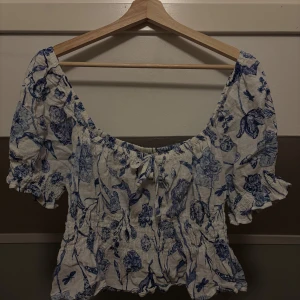 Blommig blus  - Charmig blus med blått blommönster på vit bakgrund. Den har korta ärmar och ett knytband framtill för en söt detalj. Perfekt för en somrig look!