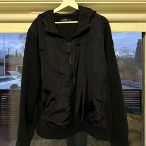 J.Lindeberg Cardigan - Snygg mörkblå cardigan från Lindberg med huva och dragkedja. Jackan har en kombination av material med en glansig framsida och stickade ärmar. Perfekt för en stilren look.