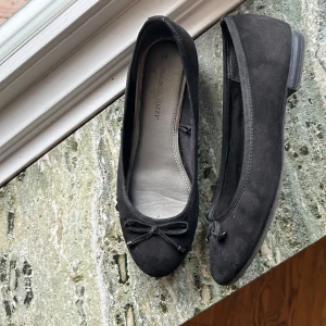 Svarta ballerinaskor från Marco Tozzi - Säljer ett par eleganta svarta ballerinaskor från Marco Tozzi. Skorna är tillverkade i mocka och har en söt rosett på tån.