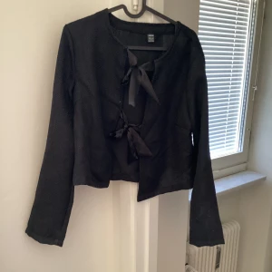 Svart kofta med rosetter Shein - Säljer en svart kavaj / kofta från Shein med knytband framtill. Jackan har en elegant design med långa ärmar och en texturerad yta. Perfekt för att ge en stilren touch till din outfit.