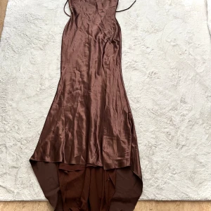 Brun axelbandslös klänning - Elegant brun axelbandslös klänning i ett glansigt material. Klänningen har en lång och flytande design med en asymmetrisk nederkant som ger en dramatisk look. Perfekt för speciella tillfällen.