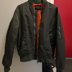  bomberjacka från Pop - Säljer en grön bomberjacka från Pop med orange foder. Jackan har en dragkedja framtill och en ficka med dragkedja på ärmen. Perfekt för en cool och avslappnad stil.