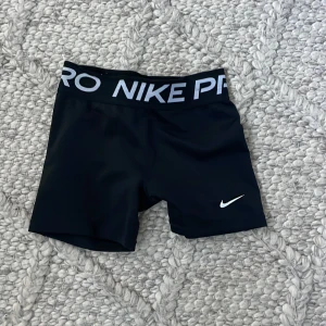 Svarta träningsshorts från Nike - Säljer ett par svarta träningsshorts i barn storlek från Nike med en elastisk midja och vit logga. Perfekta för gymmet eller löprundan. Bekväma och stilrena med en tight passform.