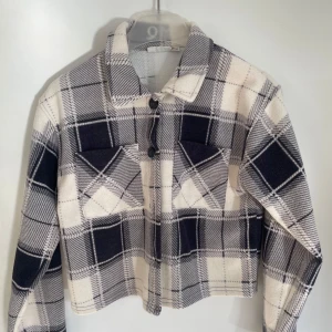 Rutig overshirt i svart och vit - Snygg rutig overshirt i svart och vit med knappar framtill. Jackan har en croppad passform och långa ärmar. Perfekt för lager-på-lager stil. Nytt skick och passar till mycket